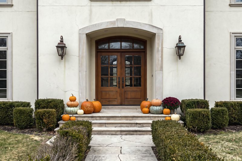 The Signature Porch on a cream stucco house with an arched entry, massed on the limestone landing either side of the glazed doors with a burgundy mum and one custom engraved pumpkin