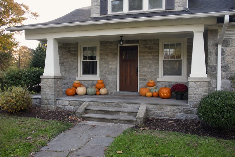 The same porch with the full Signature Porch: orange, white and green pumpkins massed on the porch floor either side of the door, a burgundy mum by the right column and one custom engraved pumpkin