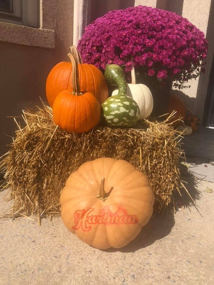 A porch display of straw bales, purple mums, and gourds with a pumpkin engraved Hartman in front