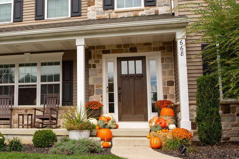 The Harvest on a stone-front colonial: pumpkins in orange, white and sage green either side of the door, a straw bale carrying an engraved house-number pumpkin, mums, mini gourds, and an engraved family-name pumpkin by the door